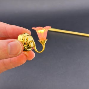 Puede incluir: Aplique de pared en miniatura de tono dorado con una pantalla de vidrio rosa. La aplique tiene un diseño decorativo y un brazo curvo.