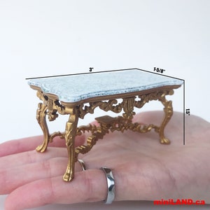 May include: Miniature gold-colored table with a marble-look top. The table is 3 inches long, 1-5/8 inches wide, and 1.5 inches high. The table has ornate, curved legs and decorative detailing. The table is a dollhouse or miniature furniture item.
