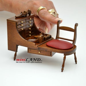 Puede incluir: Un banco de zapatero en miniatura de madera con un asiento acolchado rojo y un pequeño cajón. El banco tiene un gabinete incorporado con varios cajones y un pequeño estante para herramientas. El banco está hecho de madera oscura y tiene detalles dorados.