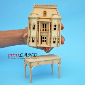 May include: A wooden dollhouse with a white paint finish and a detailed facade. The dollhouse has a front porch with a railing and a double door entrance. The dollhouse is being held in a hand above a wooden table with two drawers.