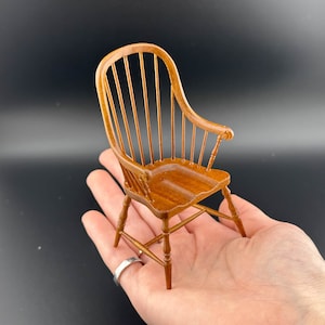 May include: A miniature wooden chair with a spindle back and a brown finish. The chair is sitting on a hand.