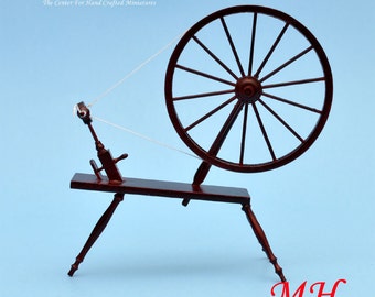 May include: A miniature wooden spinning wheel with a red finish and a white thread. The wheel has a large, spoked wheel and a smaller, crank-like handle. The spinning wheel is on a wooden base with four legs. The text "miniLAND.ca The Center for Hand Crafted Miniatures" is visible in the top left corner of the image. The text "MH" is visible in the bottom right corner of the image.