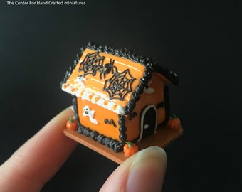 Może przedstawiać: Miniaturowy piernikowy domek na Halloween z pomarańczowymi ścianami, czarnym obramowaniem dachu i białymi detalami lukru. Dach zdobią pajęczyny i pająk. Duch i dynie zdobią dom. Tekst "miniLAND.ca". Skala 1:12.