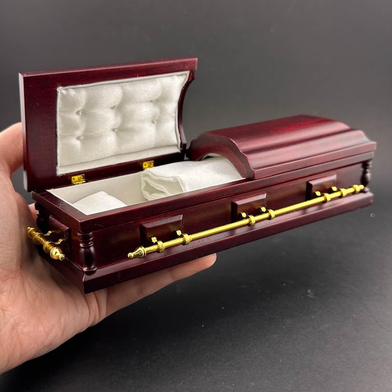 May include: A miniature wooden coffin with a gold handle and a white satin lining. The coffin is open and the lid is propped up.