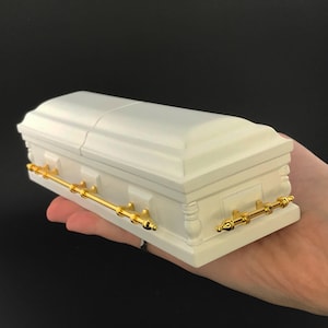 May include: A white wooden coffin with gold-colored metal handles. The coffin is closed and has a slightly rounded top.