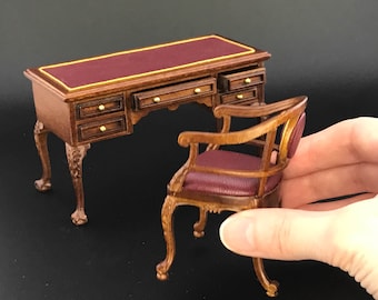 May include: A miniature wooden desk with a burgundy leather top and gold trim, featuring multiple drawers and carved legs. A matching burgundy leather chair with carved legs is included.