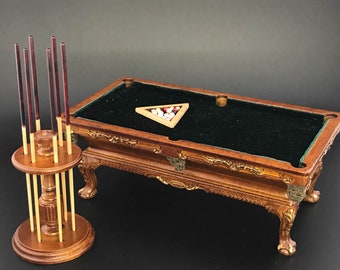 May include: Miniature wooden pool table with a green felt surface, ornate detailing, and a rack of pool cues. The table has a wooden triangle with billiard balls. The pool cues are dark red and light brown.