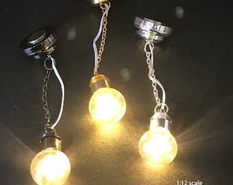 Puede incluir: Tres luminarias LED en miniatura con cadenas que cuelgan de una superficie negra. Cada luminaria tiene una base de metal de color diferente: oro, plata y negro. Las luces están encendidas y emiten un brillo blanco cálido. El texto "miniLAND.ca" está en la esquina inferior izquierda de la imagen. El texto "Escala 1:12 Luz LED a batería" está en la esquina inferior derecha de la imagen.