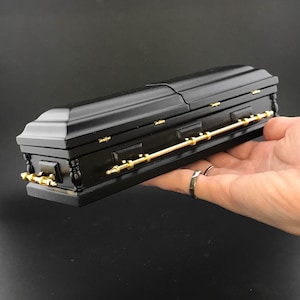 May include: A black wooden coffin with gold hinges and a gold handle. The coffin is closed and is being held in a person's hand.