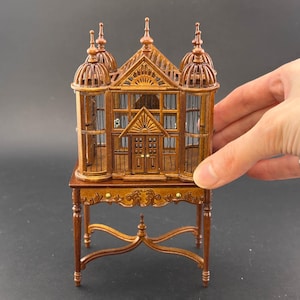 Op de afbeelding: Een miniatuur houten vogelkooi met een koepelvormige bovenkant en ingewikkelde details, geplaatst op een bijpassende houten standaard. De kooi heeft een decoratieve voordeur en is gemaakt van donkerbruin hout.