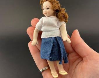 Può includere: Una bambola in miniatura con capelli ricci rossi, che indossa una camicia bianca a maniche corte e una gonna di jeans blu. La bambola ha tratti del viso dipinti e indossa scarpe gialle. La bambola è alta circa 13 cm.