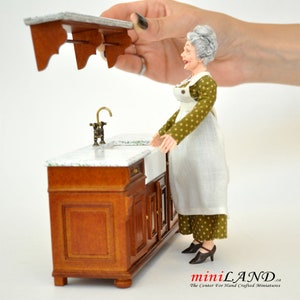 Peut inclure: Un évier de cuisine miniature pour maison de poupée avec un évier en porcelaine blanche, un robinet doré et une armoire en bois à deux portes. L'armoire a un plan de travail en marbre et une étagère en bois au-dessus de l'évier.
