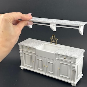 Op de afbeelding: Miniatuur witte keuken met een aanrechtblad met granietlook en een boerderijwasbak. De set bevat een bijpassend rek met een handdoekenrek. De kraan is goudkleurig. Een hand houdt de plank boven de wasbak vast.