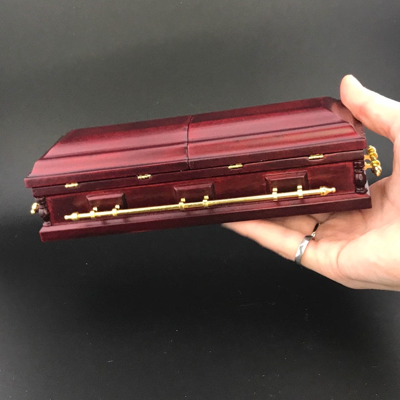 May include: A small, dark red wooden coffin with gold accents. The coffin is closed and has a hinged lid.