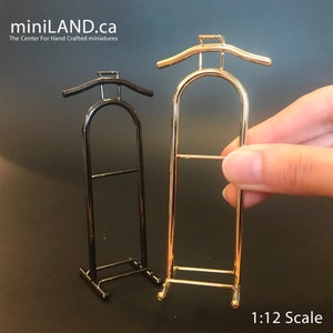Può includere: Presentazione di due mini grucce in metallo, una nera e una dorata. La gruccia dorata è tenuta da una mano. Le grucce sono in scala 1:12. Il sito web miniLAND.ca è visibile.