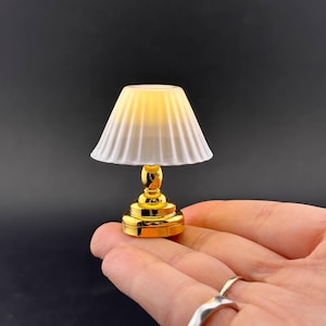 May include: A miniature gold-colored lamp with a white and yellow lampshade. The lamp has a detailed base and a fluted shade. The lamp is held in a hand against a dark background. The lamp is a decorative item.
