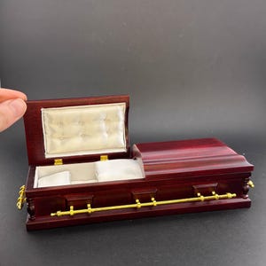 May include: A small, wooden coffin-shaped box with a gold-coloured metal handle and hinges. The lid is open, revealing a white velvet interior with two compartments.