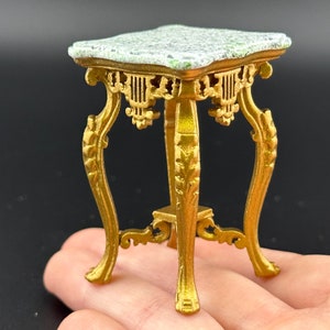 May include: Miniature gold-colored table with a square, marble-look top. The table has ornate, curved legs and decorative detailing. The tabletop has a white and green speckled pattern. A small, decorative accent piece.