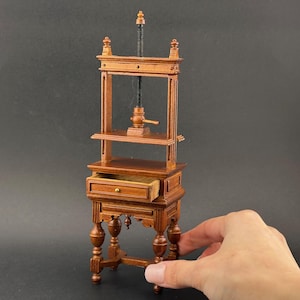 May include: Miniature wooden printing press with a drawer. The press has a dark brown finish, a screw mechanism, and ornate details. The drawer is open, revealing the interior. The press is a decorative item.