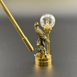Puede incluir: Un objeto decorativo de metal dorado que presenta un águila posada en una rama, con un pequeño globo de cristal y un tubo largo y delgado de metal dorado. El águila está intrincadamente detallada y el globo es transparente.