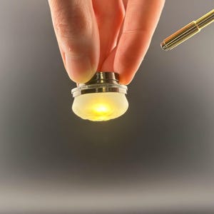 Puede incluir: Una pequeña luz redonda blanca con una base plateada. La luz está encendida y emite un brillo amarillo cálido.