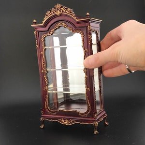 May include: A mahogany-coloured glass display cabinet with ornate gold accents and glass shelves. The cabinet has a traditional design with a curved top and intricate details.
