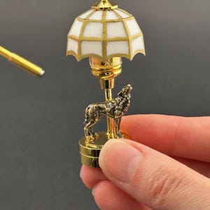 Peut inclure: Une lampe de table miniature en or avec un abat-jour à motifs blancs et dorés. La lampe est surmontée d'une figurine de loup.