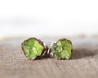 May include: A pair of earrings featuring raw green peridot stones set in copper wire. The stones are irregular in shape and have a natural, unpolished look.