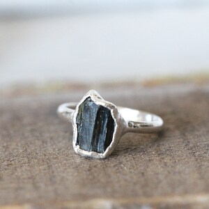 Puede incluir: Un anillo de plata con una piedra preciosa bruta de color verde oscuro y facetada. El anillo est&aacute; hecho de una banda de plata martillada.