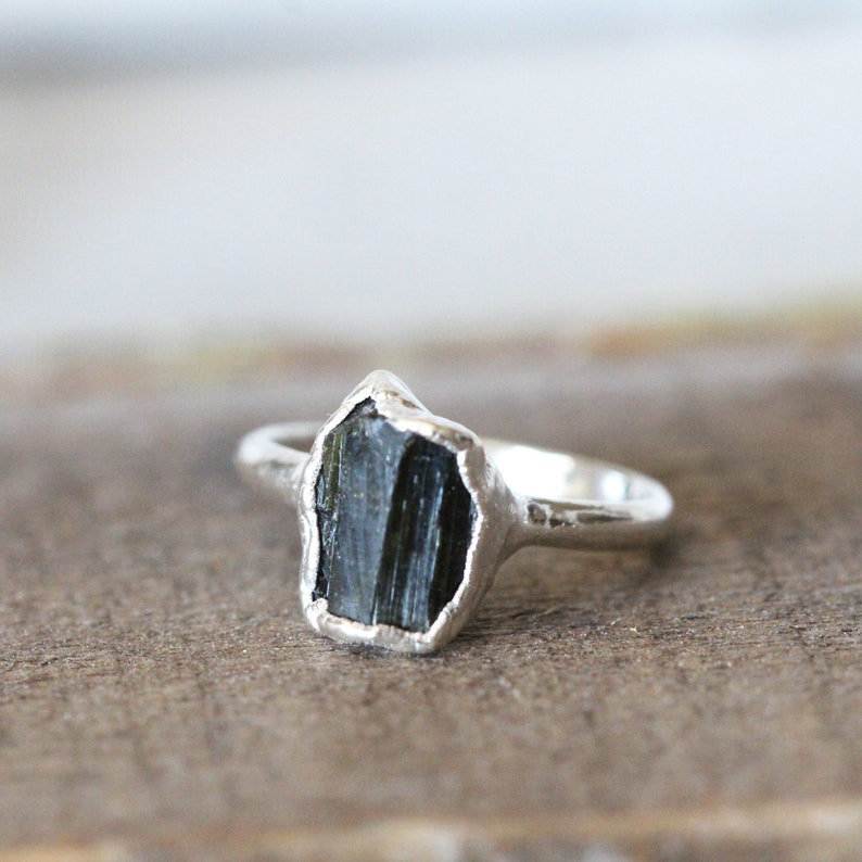 Puede incluir: Un anillo de plata con una piedra preciosa bruta de color verde oscuro y facetada. El anillo est&aacute; hecho de una banda de plata martillada.