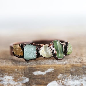 May include: A copper ring with five different colored stones set in it. The stones are a light brown, a light blue, a light yellow, a dark green, and a light green.