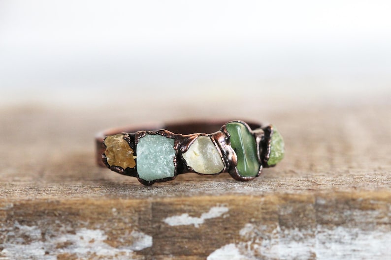 May include: A copper ring with five different colored stones set in it. The stones are a light brown, a light blue, a light yellow, a dark green, and a light green.