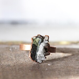 May include: A copper ring featuring a raw, uncut gemstone. The stone is a mix of dark green, black, and white crystals, set in a rustic, organic style. The ring band is a simple, polished copper.