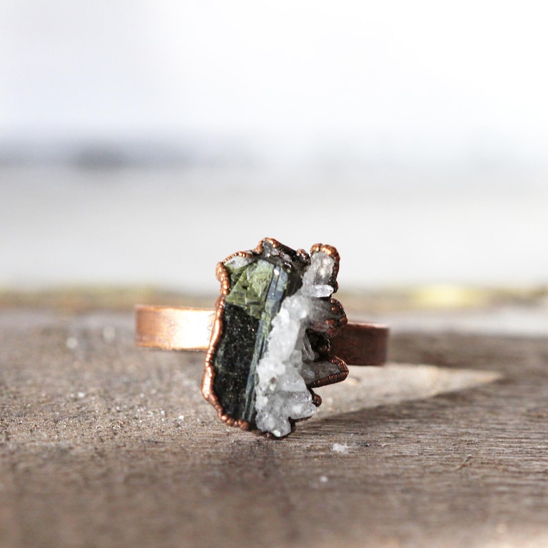 May include: A copper ring featuring a raw, uncut gemstone. The stone is a mix of dark green, black, and white crystals, set in a rustic, organic style. The ring band is a simple, polished copper.