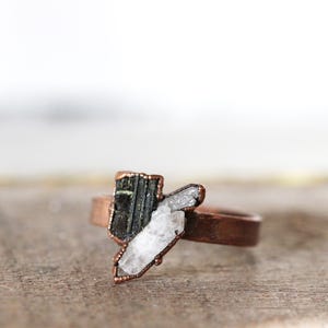 May include: A copper-colored ring featuring a dark green and white crystal design. The ring's band is a simple, smooth band. The crystals are set in a copper-colored frame, creating a unique and natural look. The ring is displayed on a wooden surface.