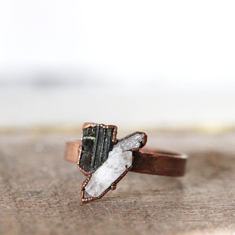 May include: A copper-colored ring featuring a dark green and white crystal design. The ring's band is a simple, smooth band. The crystals are set in a copper-colored frame, creating a unique and natural look. The ring is displayed on a wooden surface.