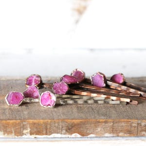 May include: A collection of hair pins with raw, pink gemstone accents. The gemstones are set in silver-colored metal and are displayed on a wooden surface. The hair pins have a natural, organic aesthetic.