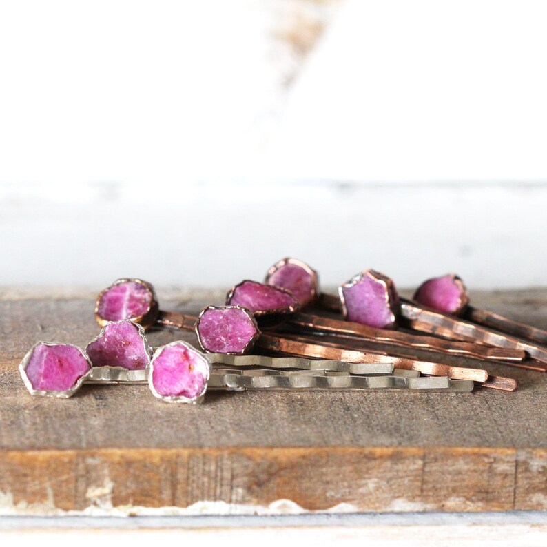 May include: A collection of hair pins with raw, pink gemstone accents. The gemstones are set in silver-colored metal and are displayed on a wooden surface. The hair pins have a natural, organic aesthetic.
