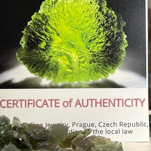 May include: A close-up of a certificate of authenticity for green Moldavite crystals. The certificate is printed on white cardstock with black text. The text reads "CERTIFICATE of AUTHENTICITY Fine Jewelry, Prague, Czech Republic, according to the local law". The image also shows a pile of green Moldavite crystals.