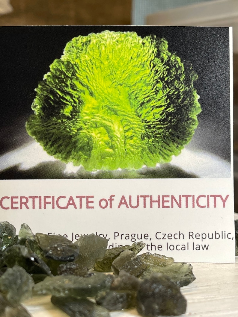 May include: A close-up of a certificate of authenticity for green Moldavite crystals. The certificate is printed on white cardstock with black text. The text reads "CERTIFICATE of AUTHENTICITY Fine Jewelry, Prague, Czech Republic, according to the local law". The image also shows a pile of green Moldavite crystals.