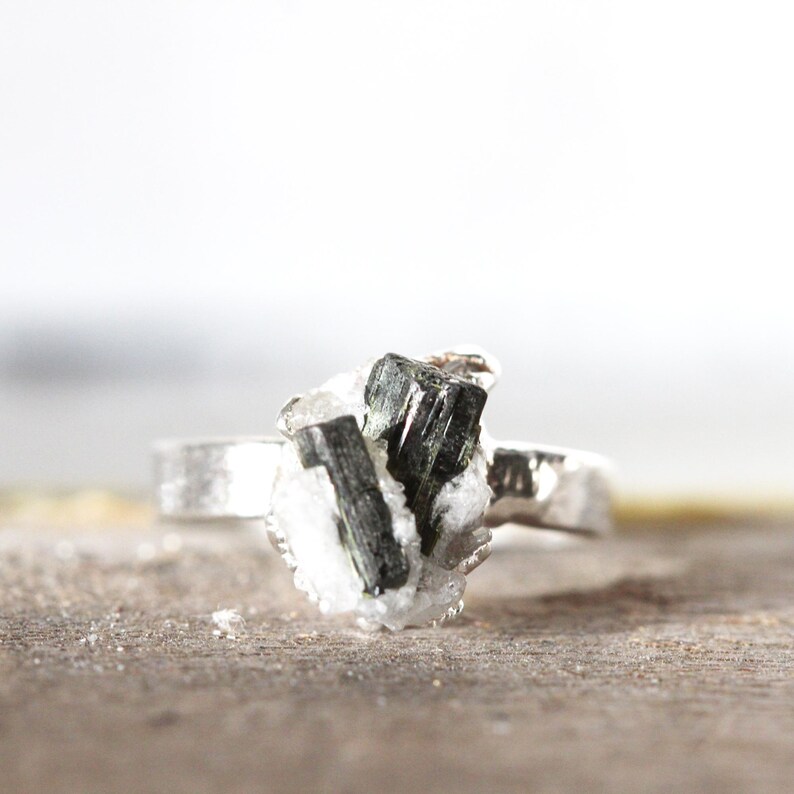 May include: A silver ring featuring a cluster of dark green tourmaline crystals set in a white matrix. The ring has a simple, textured band and a natural, organic aesthetic. The gemstone cluster measures approximately 0.75 inches in length.