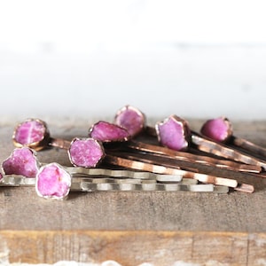 May include: A collection of hair pins with raw, pink gemstones set in copper and silver metal. The gemstones have a rough, natural texture. The pins are arranged on a wooden surface, with a blurred white background.