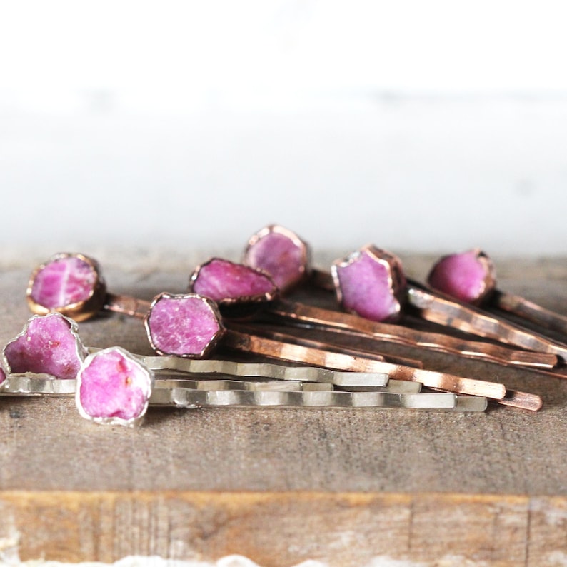 May include: A collection of hair pins with raw, pink gemstones set in copper and silver metal. The gemstones have a rough, natural texture. The pins are arranged on a wooden surface, with a blurred white background.