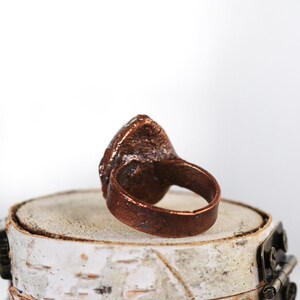 May include: A copper ring with a large, irregular, light blue stone set in the center. The ring is sitting on a small, white, wooden platform.