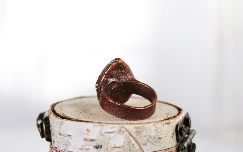 May include: A copper ring with a large, irregular, light blue stone set in the center. The ring is sitting on a small, white, wooden platform.