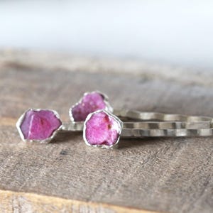 May include: Three silver rings with raw, pink ruby stones. The rings are set on a wooden surface. The rubies have a natural, irregular shape and are encased in a silver setting.