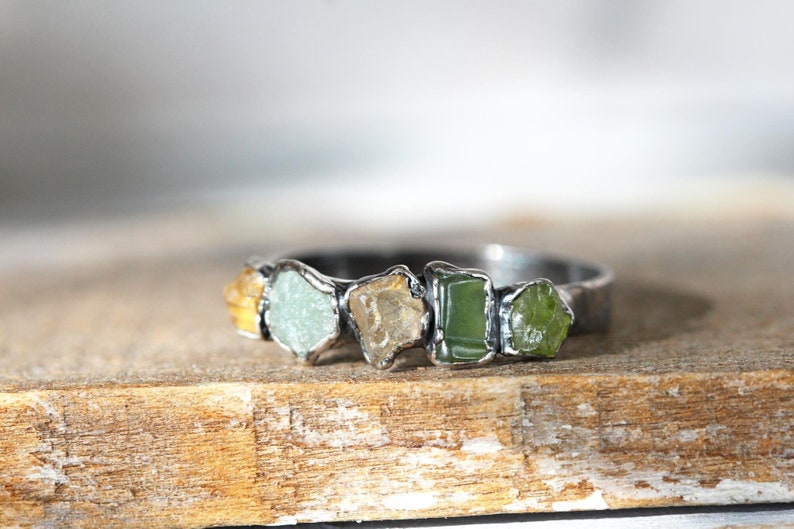 May include: A silver ring with five raw gemstones set in the band. The stones are a mix of yellow, green, and light green.
