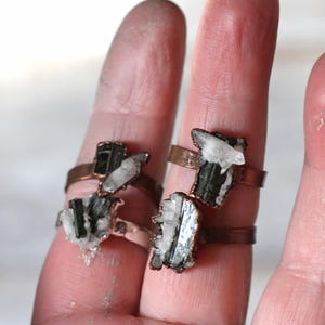 May include: Four copper-colored rings, each featuring a unique combination of black and white crystals. The crystals are irregularly shaped and mounted on simple, adjustable bands. The rings are displayed on fingers against a soft, neutral background.