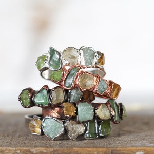 May include: Stack of four rings made with raw, uncut gemstones. The rings are made with copper and silver wire and feature green, yellow, and clear stones.