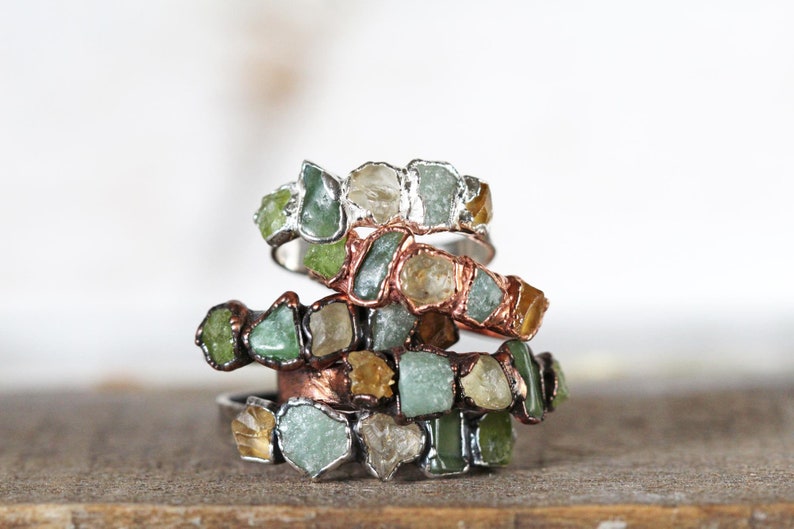 May include: Stack of four rings made with raw, uncut gemstones. The rings are made with copper and silver wire and feature green, yellow, and clear stones.